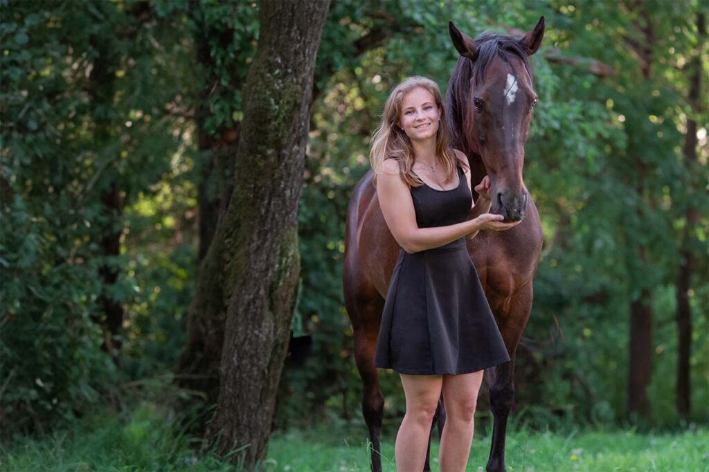 Blondes Fotomodelin einem schwarzen Minikleid posiert bei einem Pferdefotoshooting mit einem braunen Pferd.