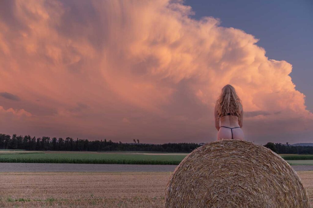 Blondes Fotomodel sitzt in sexy Dessous/Lingerie auf einem Strohballen und schaut im Sonnenuntergang einer Gewitterwolke zu.