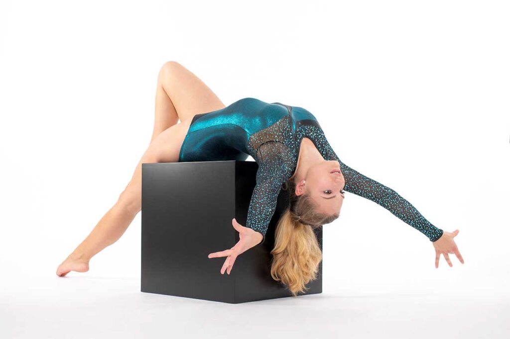 Blondes Sport-Geräteturnen- Kunstturnen Fotomodel posiert in einem blau glänzenden Leotard auf einem schwarzen Würfel im Fotostudio bei einem Fotoshooting.